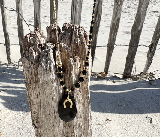 Talisman chic et noir intemporel.
Collier sur chaîne maille fantaisie en or laminé - Pendentif "goutte" en onyx noire et petite breloque fer à cheval en or laminé - Perles d'onyx et obsidienne et en or laminé 14 carats.